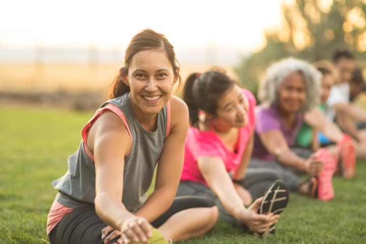 Women sitting on grass in a park, stretching and smiling during a group exercise or yoga class, with trees and sunlight in the background.