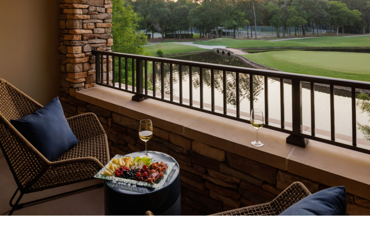 Two chairs on a balcony overlook a golf course, with a platter of food and two glasses of white wine on a small table and the balcony ledge.