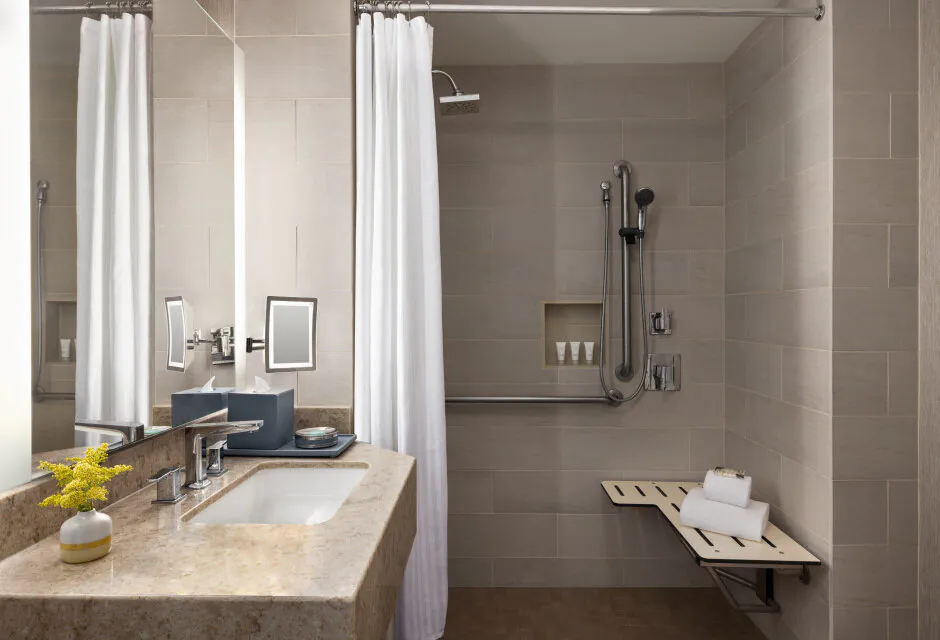 Accessible bathroom with beige tiles, a roll-in shower, white curtain, grab bars, wall bench, and vanity with sink, mirror, and toiletries.