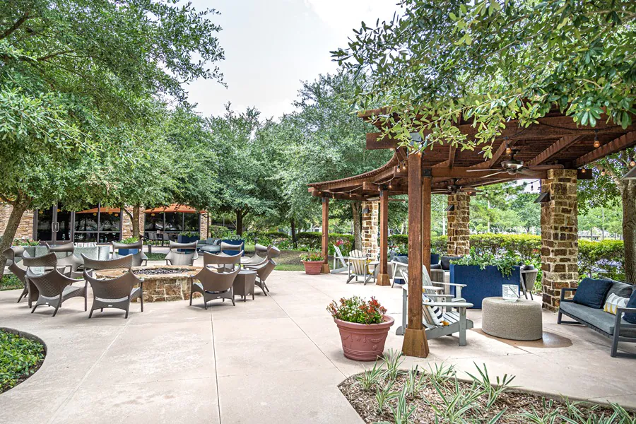 Outdoor patio with tables, chairs, and couches under wooden pergolas, surrounded by lush green trees and potted plants.