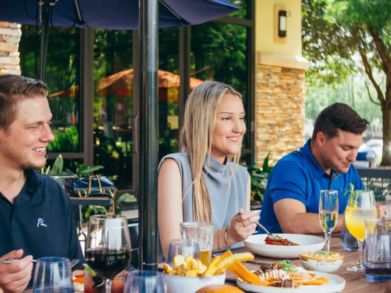 Three people sit at an outdoor restaurant table, eating and drinking together, with plates of food and drinks in front of them on a sunny day.