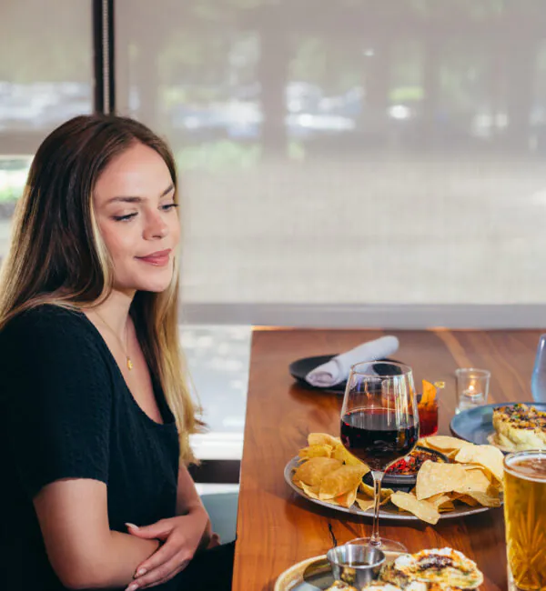Woman sitting at a table with wine, chips, oysters, and other food, looking down and smiling in a restaurant setting.