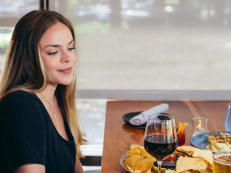 Woman sitting at a table with wine, chips, oysters, and other food, looking down and smiling in a restaurant setting.