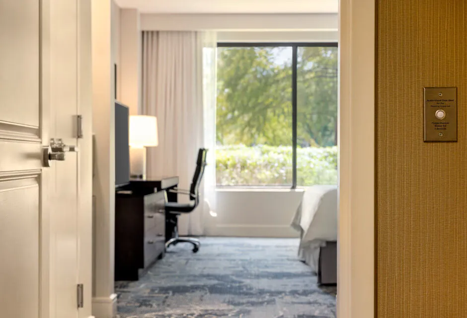 A modern hotel room with a bed, desk, office chair, lamp, and large window with trees visible outside.