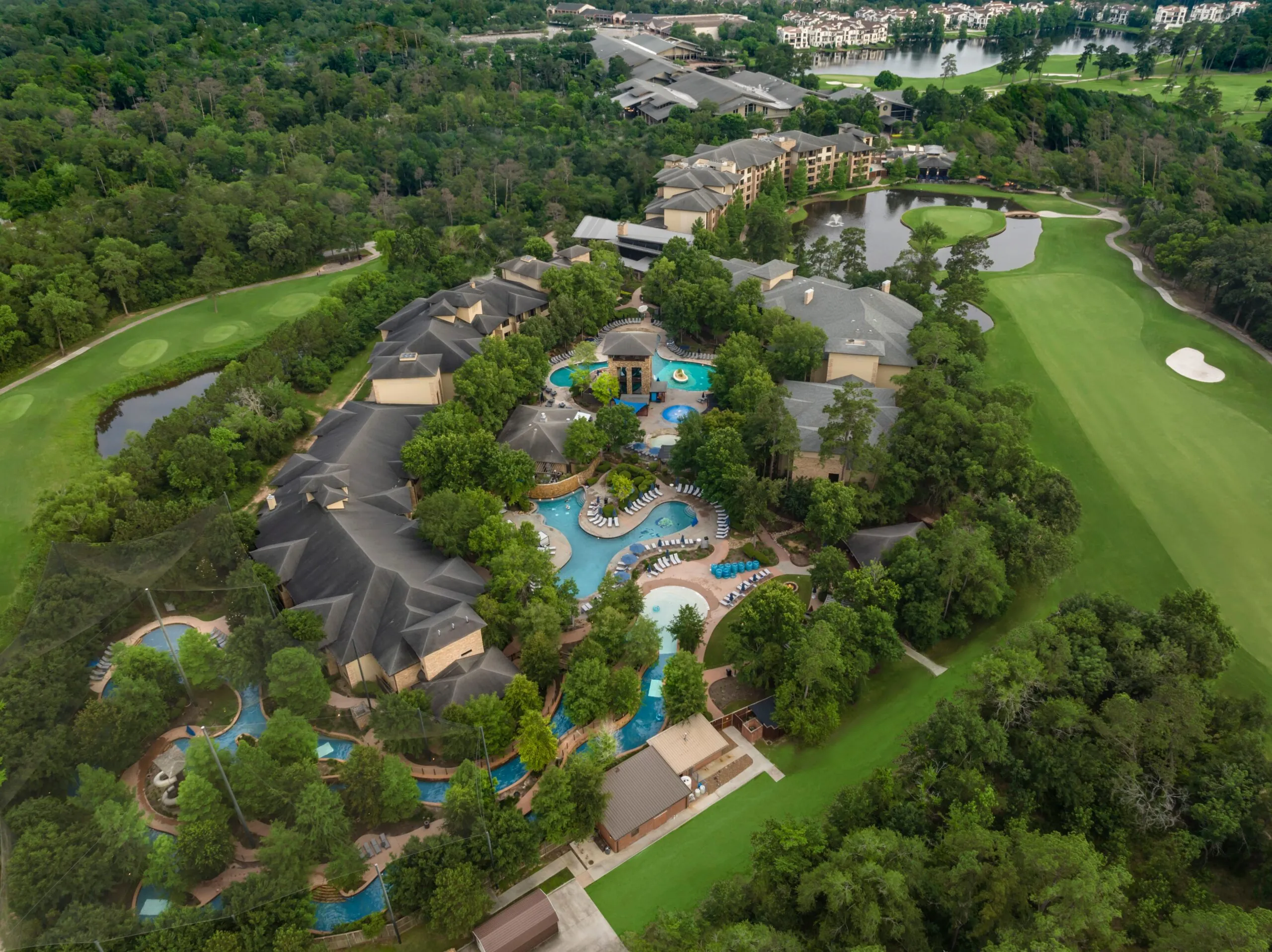Aerial view of a resort with multiple pools, surrounded by trees and next to a golf course. Buildings and lounge chairs are scattered throughout.