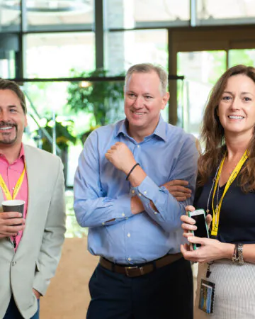 Three people with conference lanyards stand indoors, smiling. One holds a coffee cup, another a phone. Bright lobby with natural light in background.