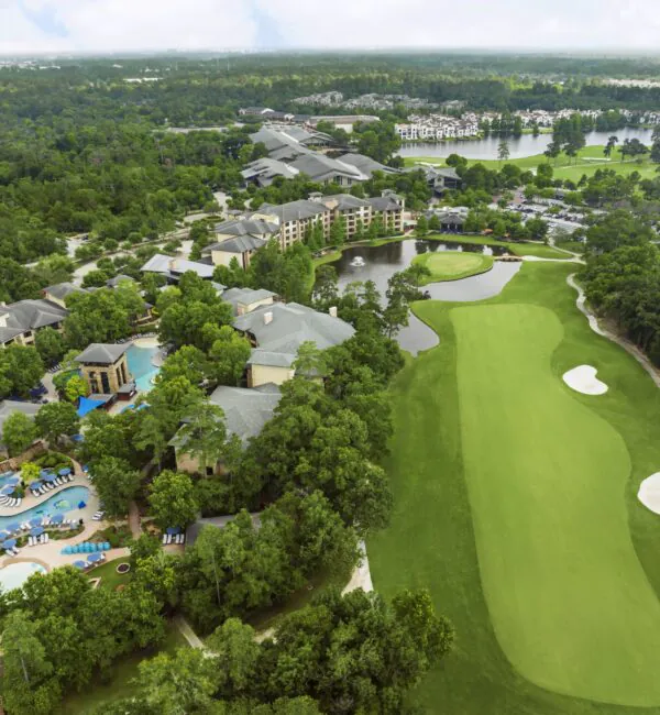 Aerial view of a large resort with pools, green golf course, sand bunkers, trees, and nearby buildings surrounded by forest.