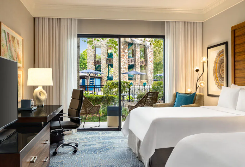 Hotel room with two beds, desk, chair, TV, and a large window showing a view of a pool area with greenery and umbrellas outside.