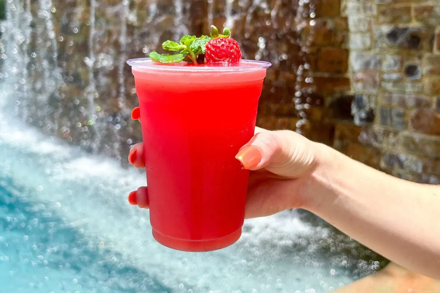 A hand holds a bright red drink with a strawberry and mint garnish by a pool and waterfall.