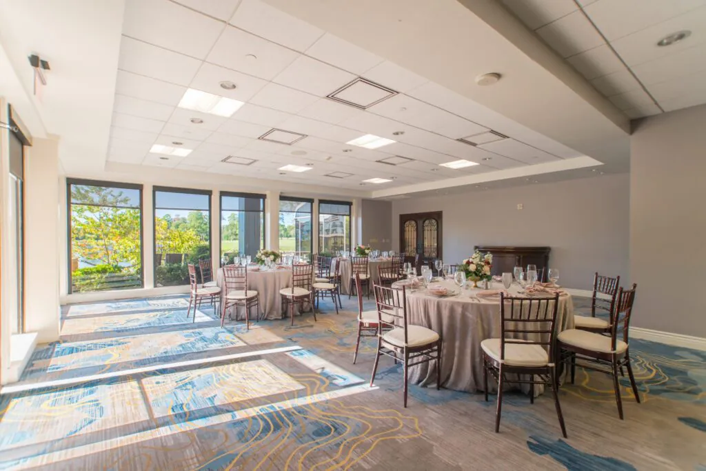 Bright banquet room with round tables set for an event, surrounded by large windows letting in natural light.