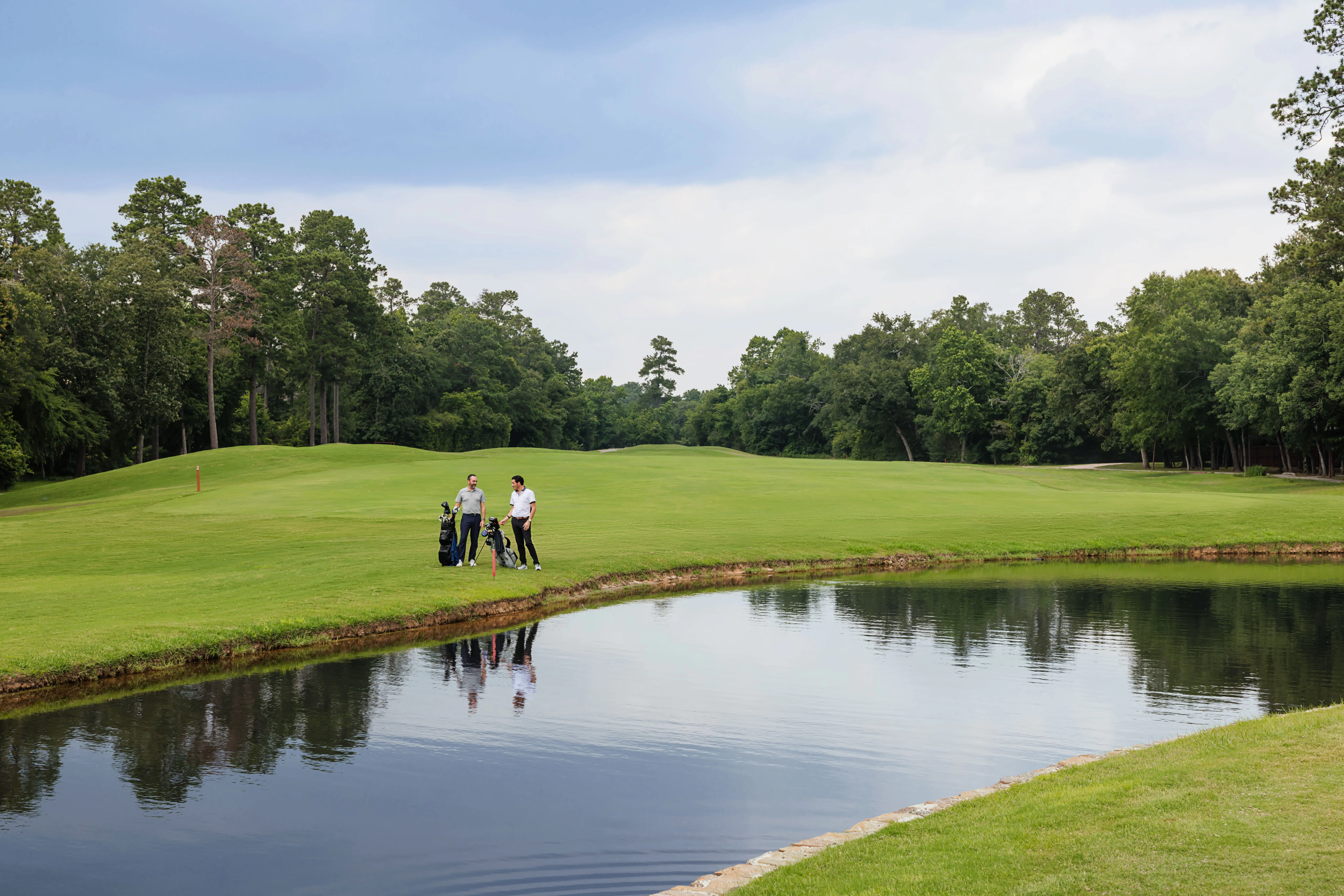Three people with golf bags stand by a pond on a green golf course, surrounded by trees under a cloudy sky.
