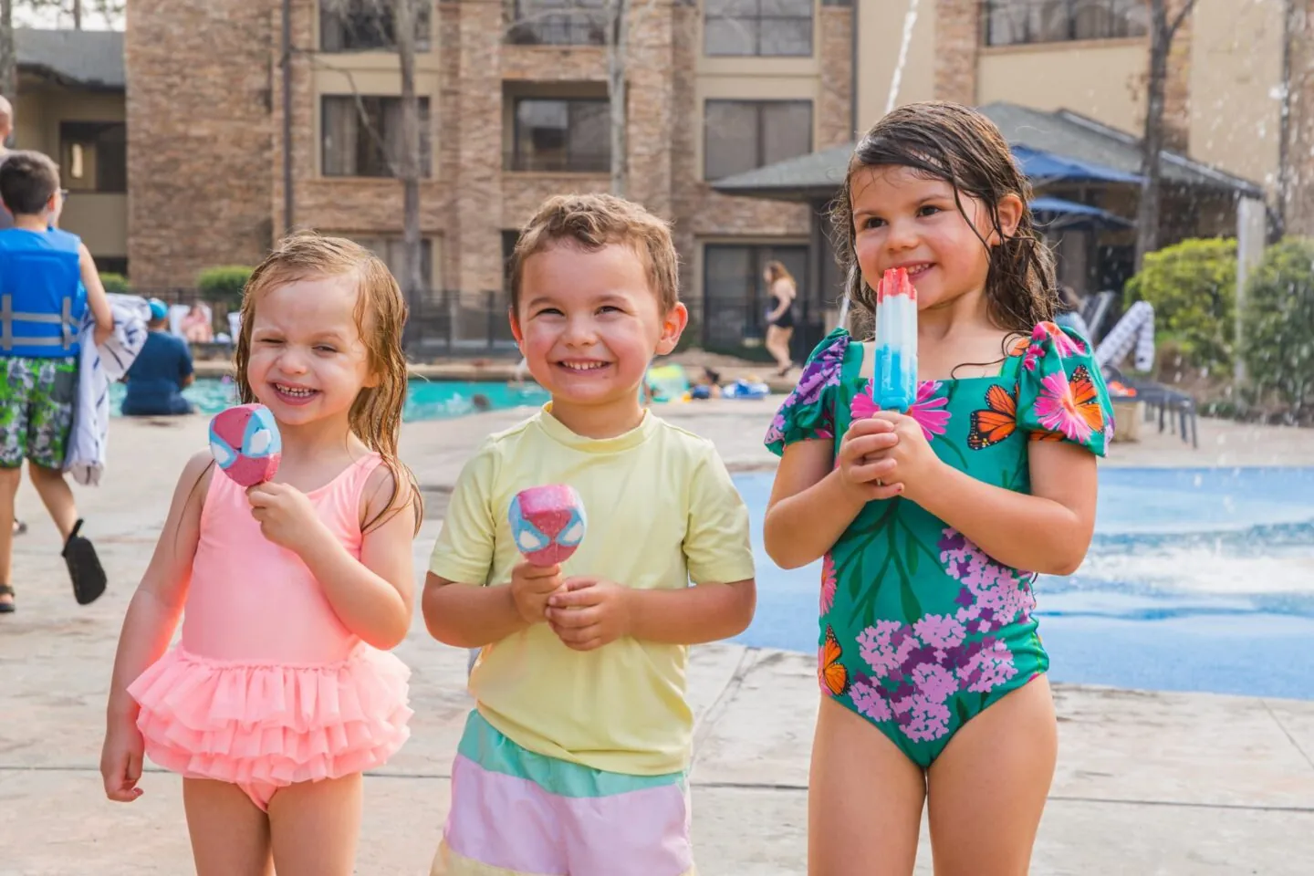 Three young children in swimsuits smile and hold popsicles by a pool on a sunny day, with water features and a building in the background.