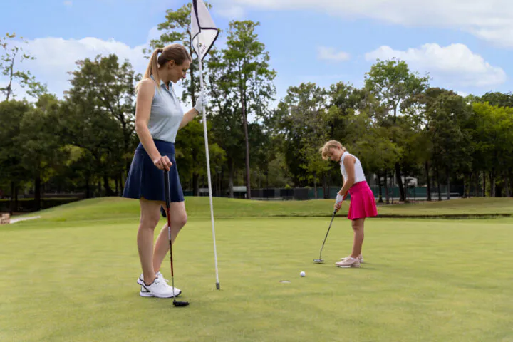 Two girls on a golf green: one holds a flagstick while the other putts a golf ball toward the hole on a sunny day with trees in the background.