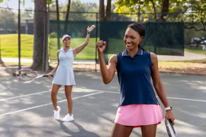 Two women on a tennis court celebrate; one smiles and pumps her fist in the foreground, the other waves her racket in the background.