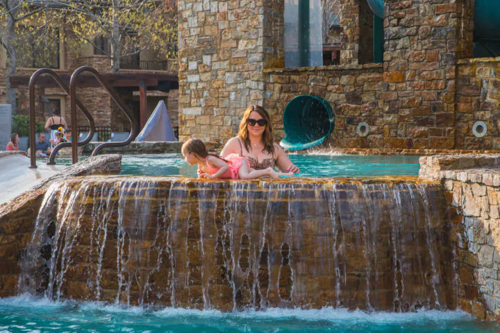 A woman and a girl relax on the edge of a stone waterfall in a pool, with water flowing over the ledge and a waterslide in the background.