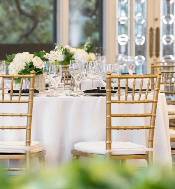 A round table with white linens, gold chairs, floral centerpieces, and set glassware in an elegant, sunlit event space.