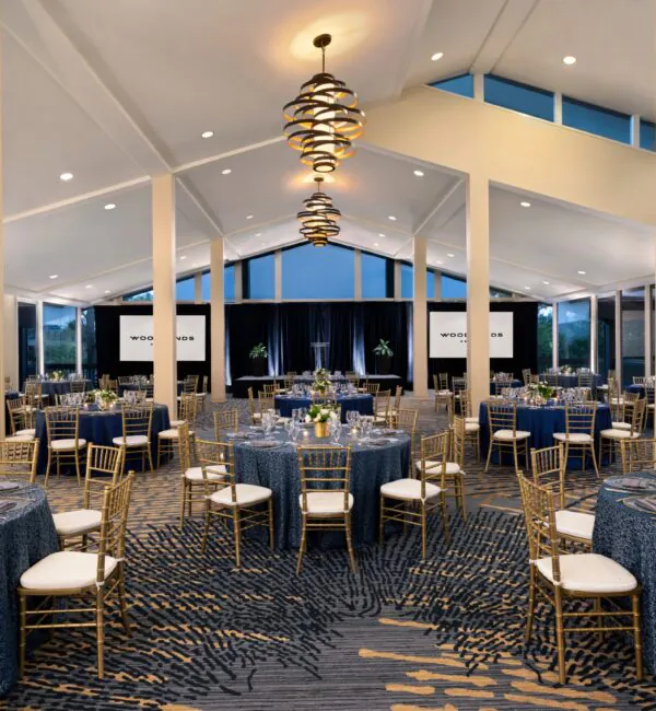 Elegant banquet hall with blue tablecloths, gold chairs, modern chandeliers, and large windows, set up for a formal event or wedding reception.