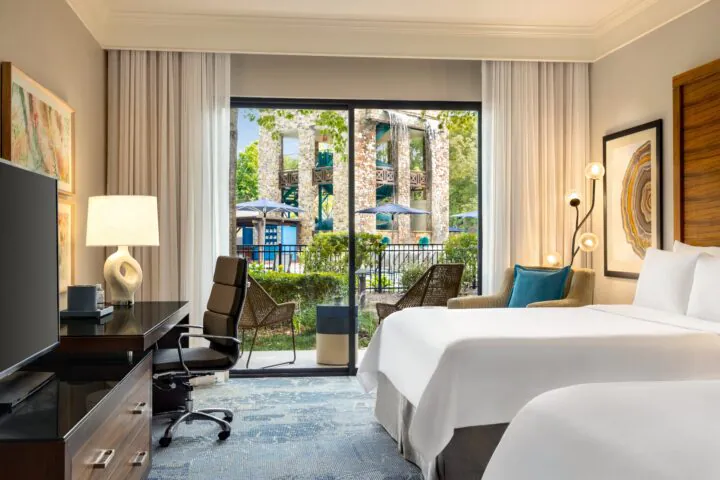 Modern hotel room with a desk, TV, armchairs, and a bed, featuring large windows overlooking a garden and outdoor pool area.