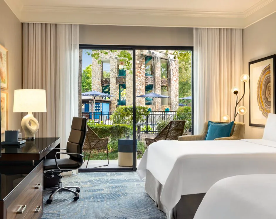 Modern hotel room with a desk, TV, armchairs, and a bed, featuring large windows overlooking a garden and outdoor pool area.