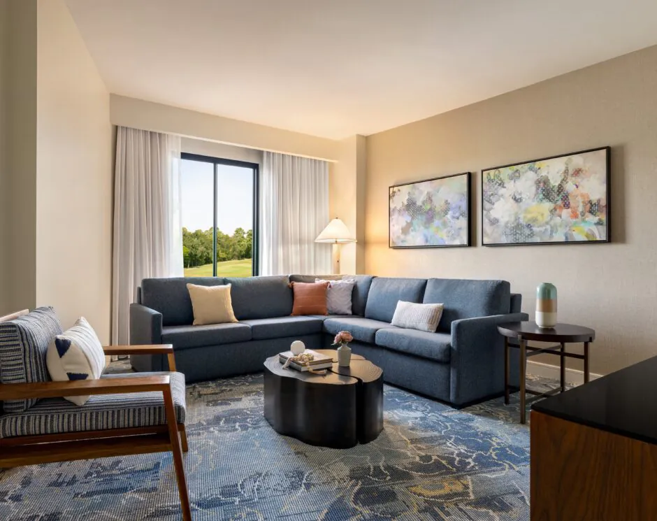 Modern living room with a gray sectional sofa, coffee table, armchair, two abstract paintings, and a window with a view of greenery.
