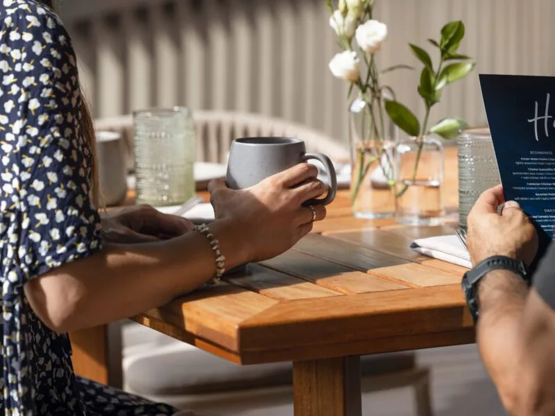 Two people sit at a wooden table; one holds a gray mug, the other holds a menu. White flowers in glass vases are on the table.