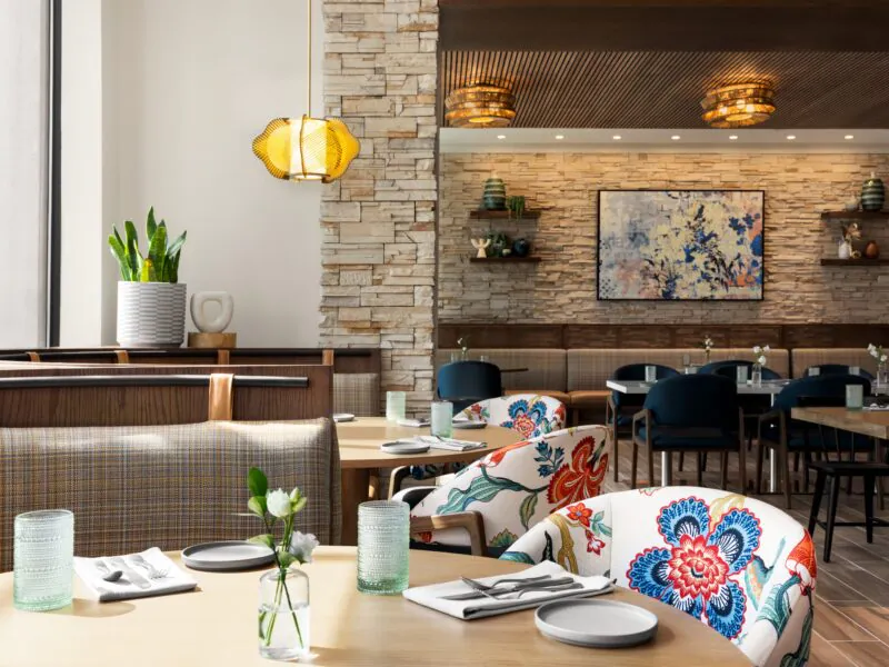 Bright restaurant with floral chairs, set tables, stone walls, hanging lights, and modern decor, with plants and artwork adding color.
