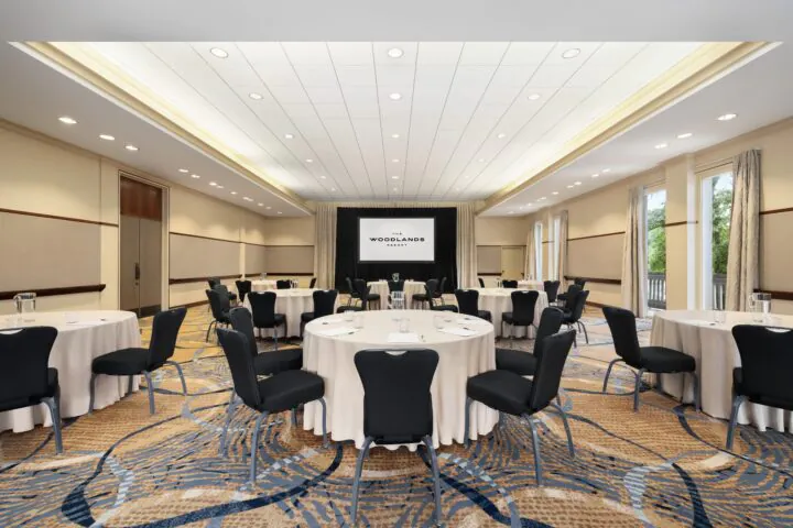 A conference room with round tables, black chairs, notepads, water glasses, and a screen at the front, lit by ceiling lights and natural window light.