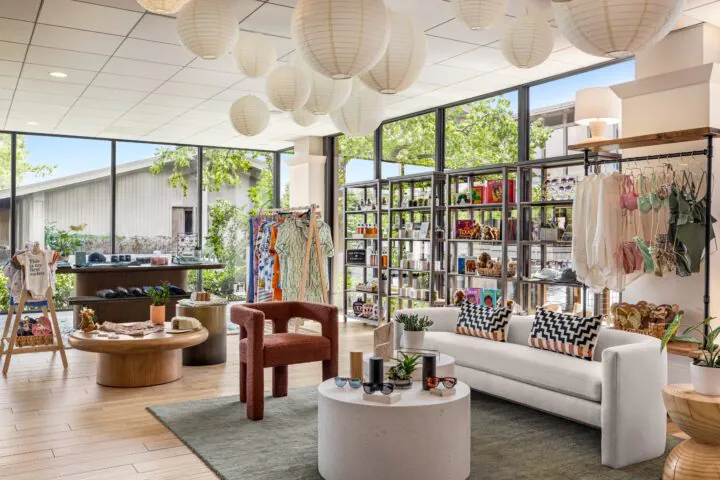 Modern boutique with cozy seating, hanging lanterns, clothing racks, shelves of goods, and large windows letting in natural light.