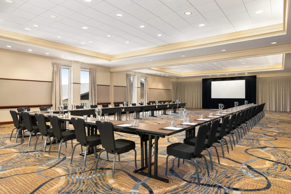 A conference room with U-shaped tables, chairs, notepads, water glasses, and a projector screen at the front.