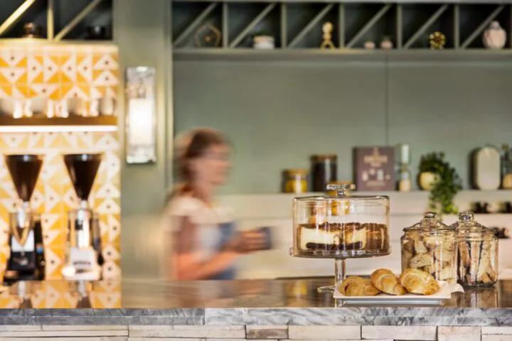 A blurred person with a mug stands behind a café counter displaying cake, cookies in jars, and pastries on a plate.