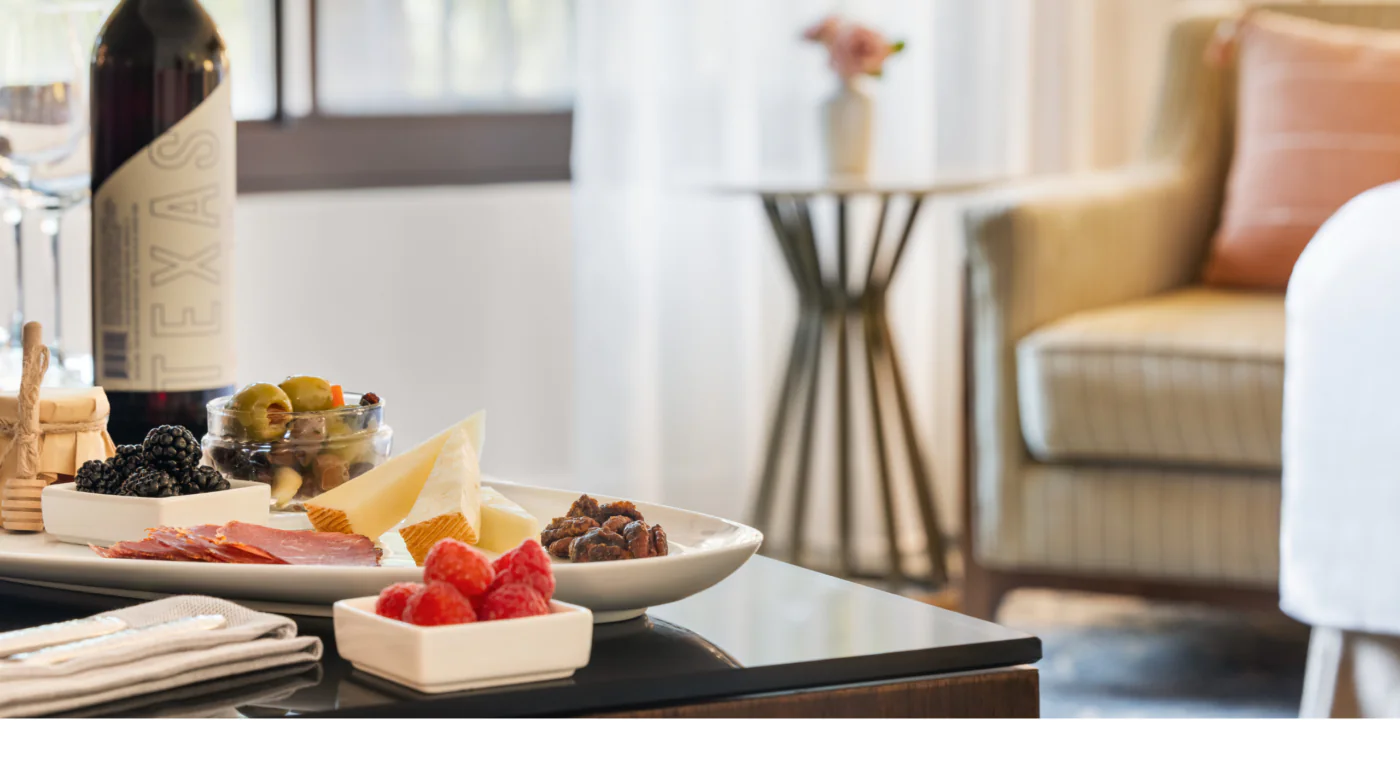 A plate of cheese, fruit, and nuts sits on a table with a wine bottle nearby; a cozy armchair and side table are in the background.