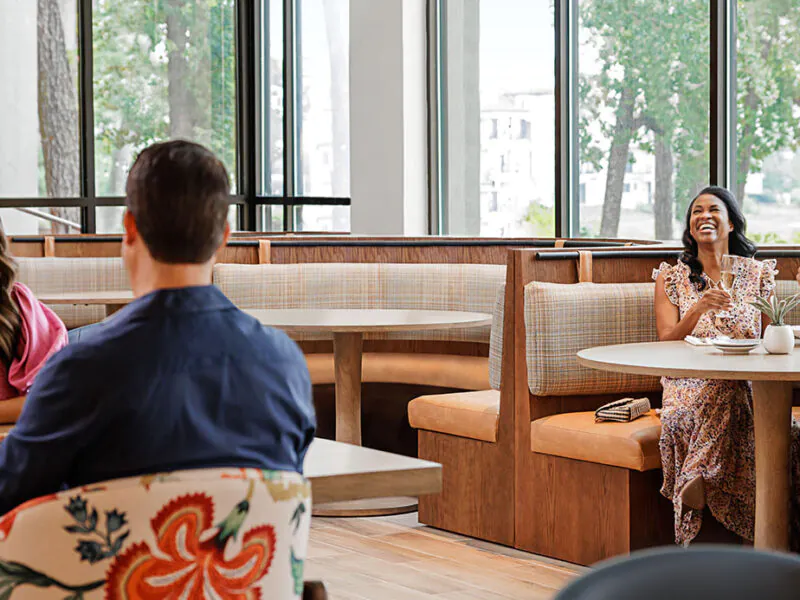 Two couples sit at separate tables in a bright restaurant, smiling, talking, and enjoying drinks and food near large windows with a view of trees.