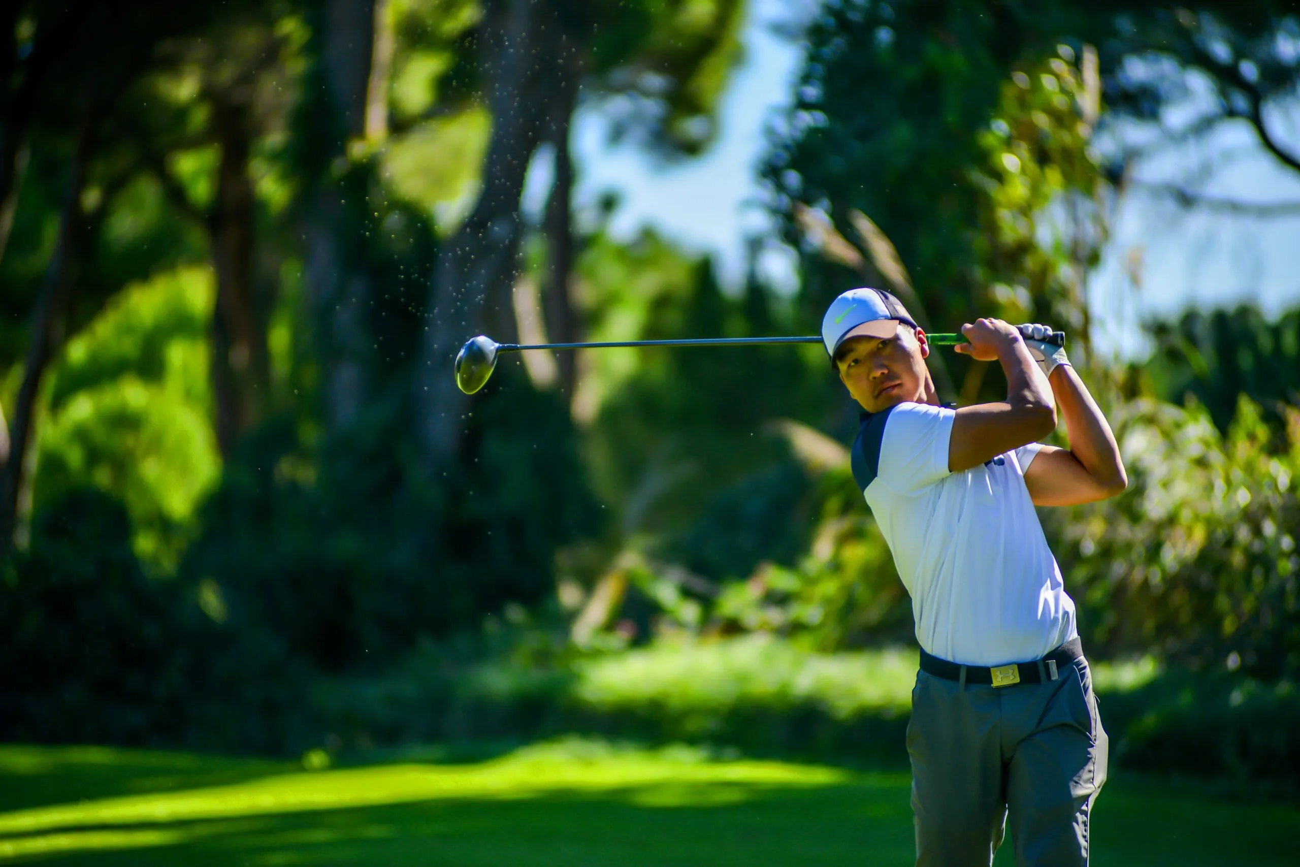 A golfer wearing a white shirt and cap swings a club on a bright, sunny course surrounded by green trees.