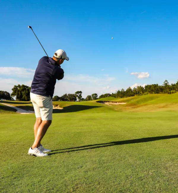 A golfer in shorts and a cap swings a club on a sunny golf course, sending the ball flying across the green fairway.