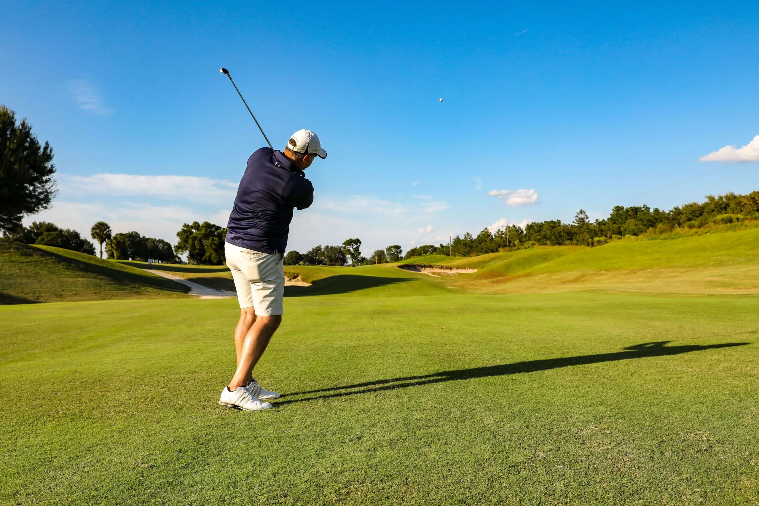 A golfer in shorts and a cap swings a club on a sunny golf course, sending the ball flying across the green fairway.