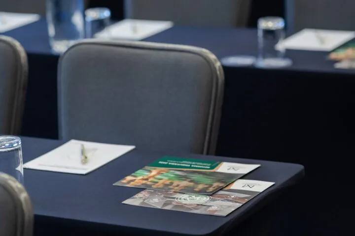 Rows of gray chairs and black tables set up for a conference, with water bottles, glasses, and papers placed on each table.