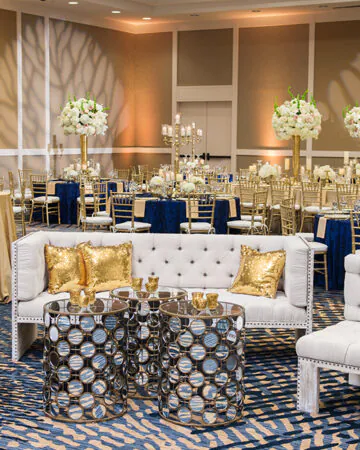 Elegant banquet hall with gold and navy decor, round tables, tall white floral centerpieces, and a plush white lounge seating area in the foreground.
