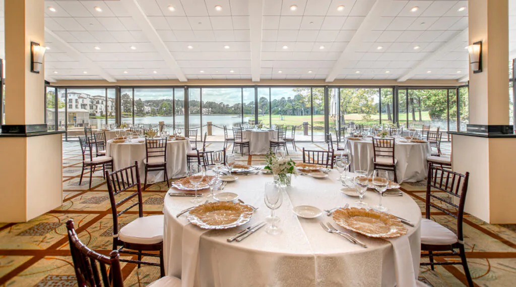 Elegant banquet hall with round tables set for a formal event, large windows overlooking a lake and green landscape, natural light filling the room.