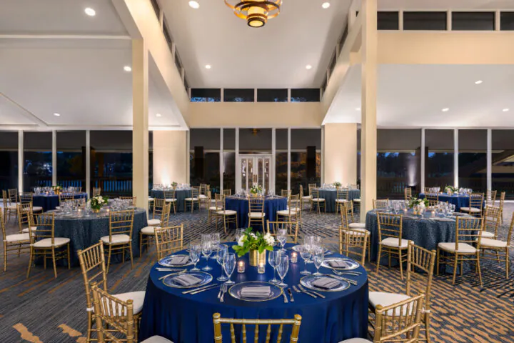 Elegant banquet hall with round tables, navy blue tablecloths, gold chairs, and floral centerpieces set for an event. Large windows line the walls.