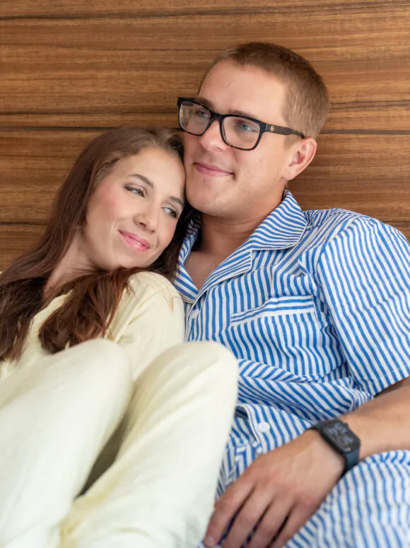 A couple in pajamas sits on a white bed, leaning against a wooden headboard, smiling and looking content.