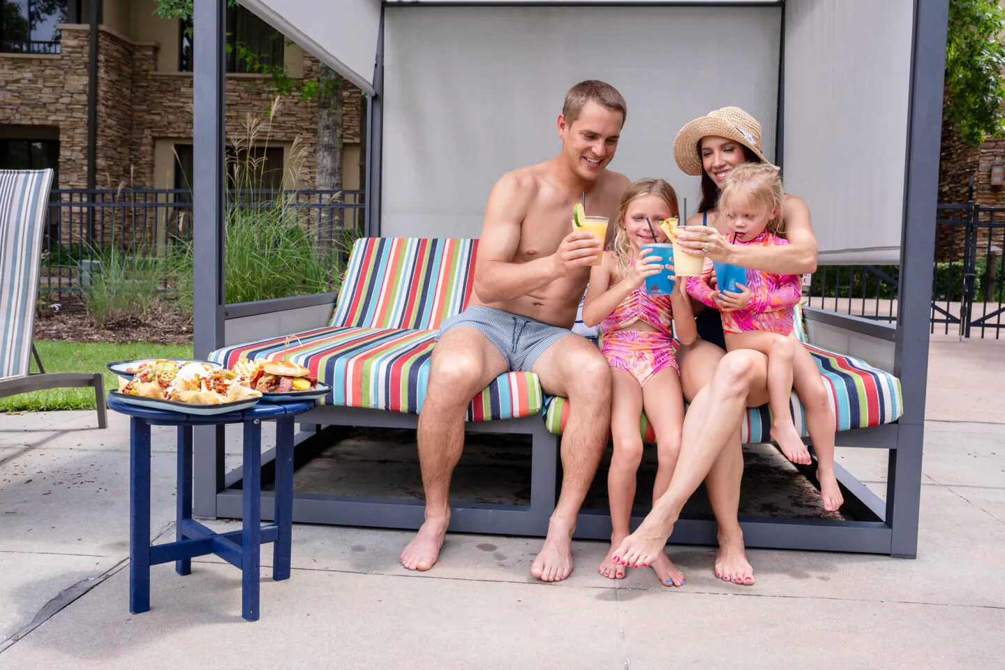 A smiling family of four in swimsuits sits on a striped poolside lounge, enjoying drinks together. Food is on a small table beside them.