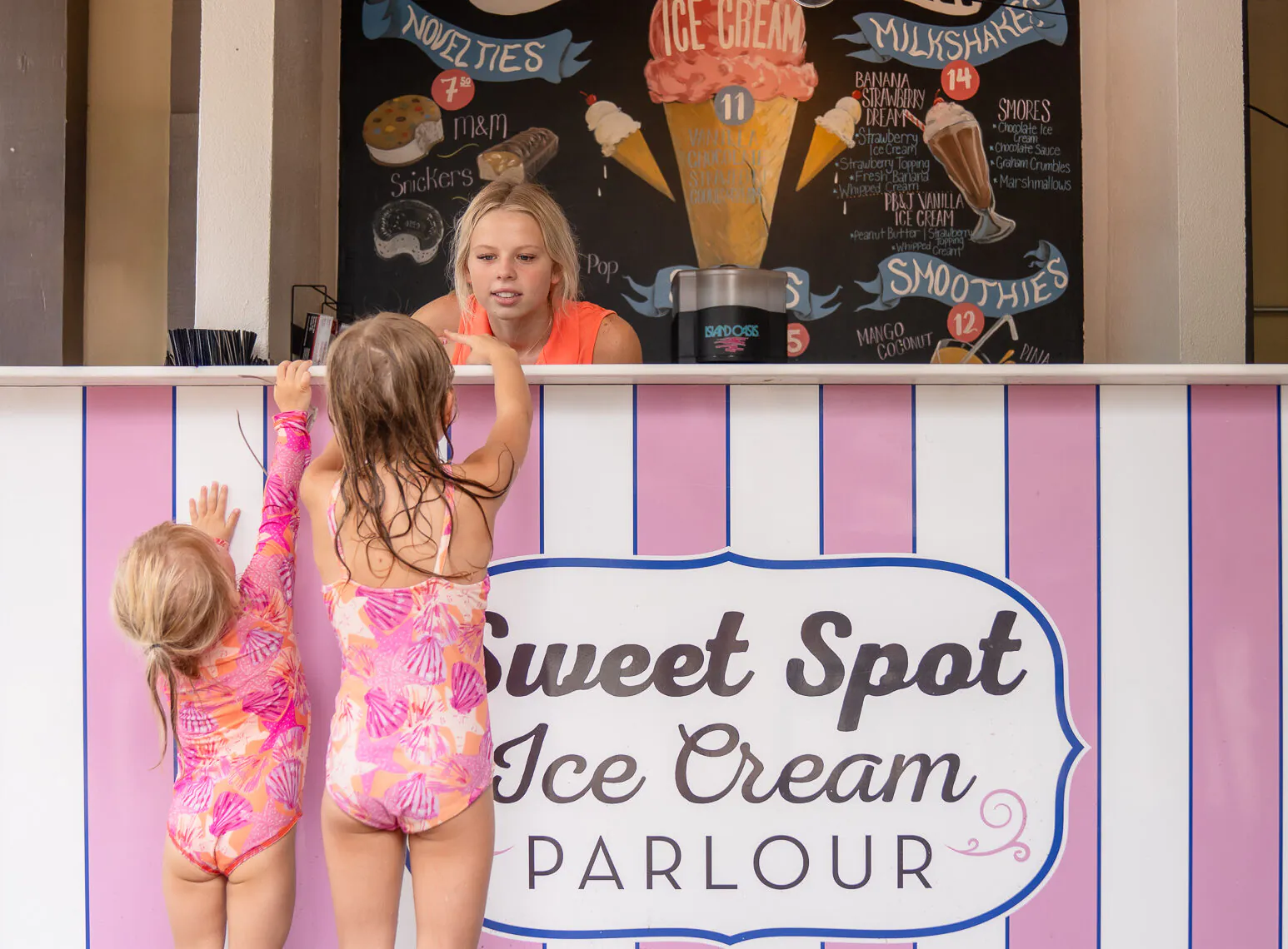 Two young girls in swimsuits reach up at an ice cream stand, where a woman is serving at the Sweet Spot Ice Cream Parlour counter.