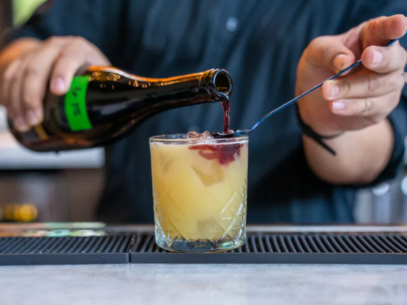 A person pours a dark liquid from a bottle into a yellow cocktail with ice, using a bar spoon, on a counter.