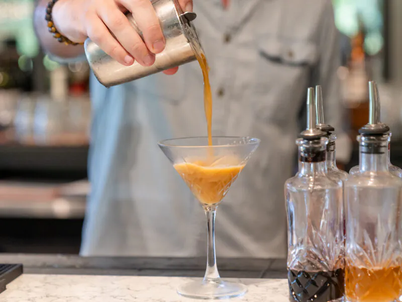 A bartender pours a brown cocktail from a shaker into a martini glass at a bar, with bottles and lime slices visible nearby.