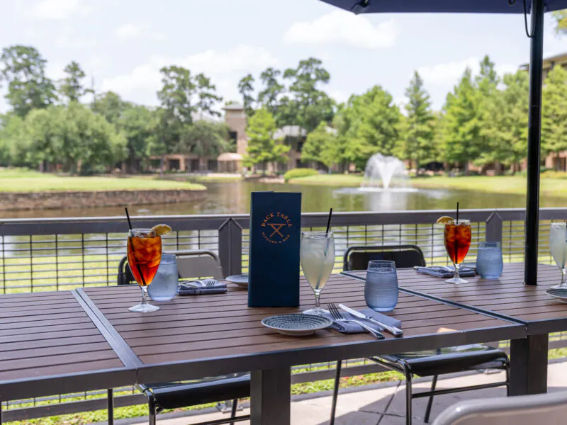 Outdoor patio table set with drinks, plates, and a menu, overlooking a pond and fountain surrounded by trees on a sunny day.