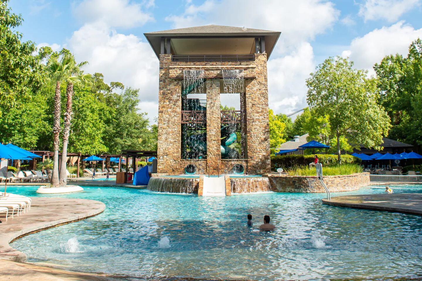 A large stone water tower with cascades overlooks a pool with people swimming, surrounded by trees and blue umbrellas on a sunny day.
