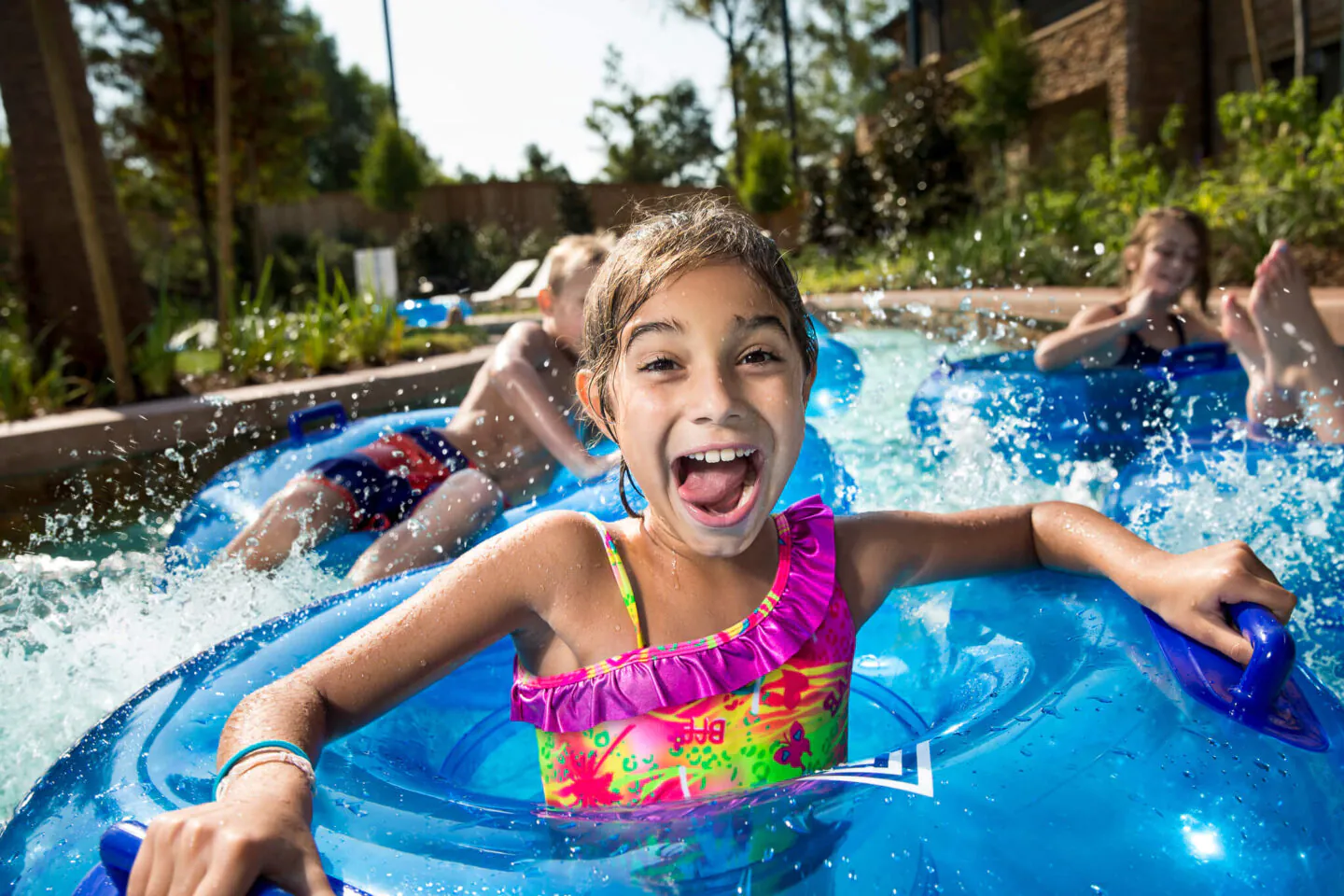 Smiling child in a colorful swimsuit sits in a blue inner tube at a water park, with water splashing and other kids in the background.