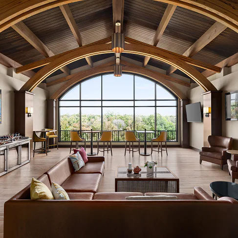 Spacious lounge with leather sofas, large arched window, wooden ceiling beams, and a view of trees outside. Coffee tables and chairs are arranged neatly.