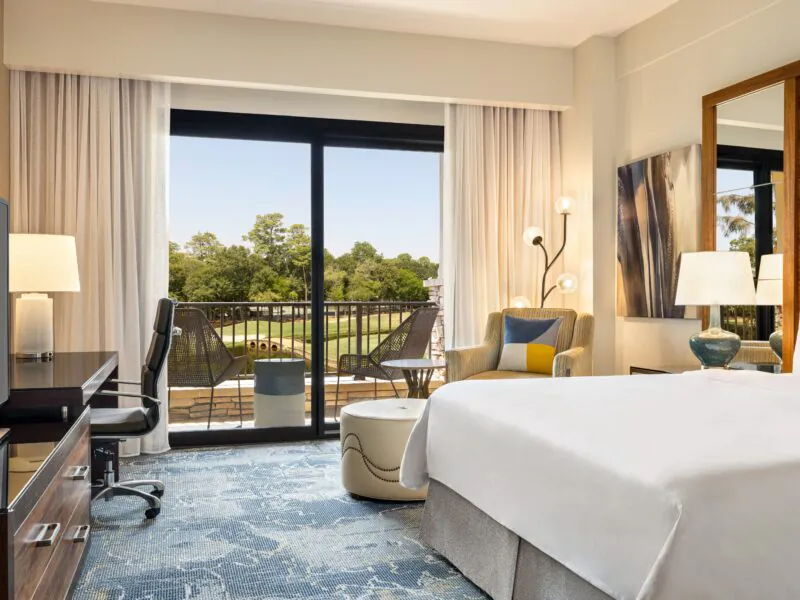Modern hotel room with a bed, desk, TV, armchair, and balcony with chairs overlooking a green outdoor view through large glass doors.