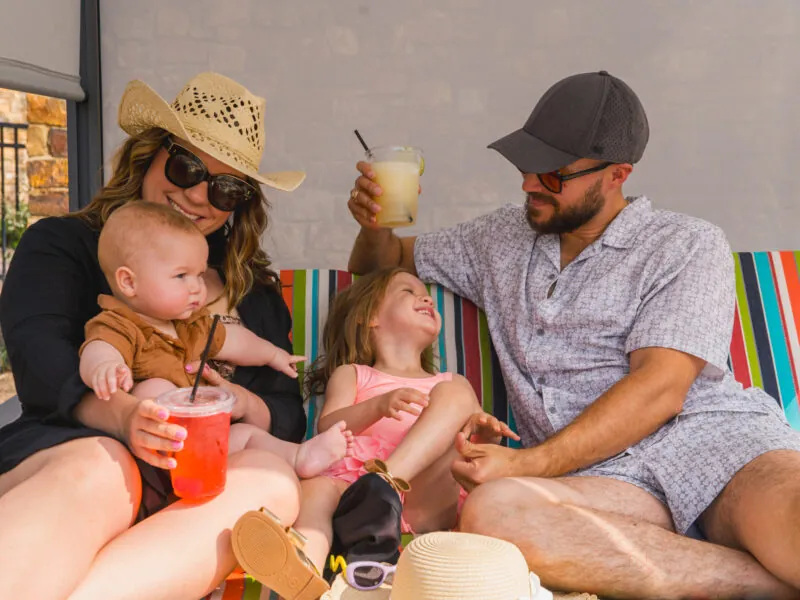 A family of four relaxes on a colorful striped lounge chair, smiling and enjoying drinks together outdoors.